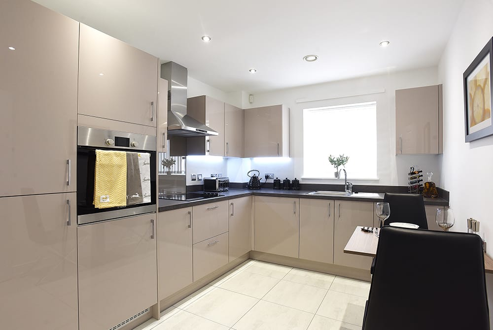 Modern fitted kitchen with beige cabinets, integrated oven and stainless steel extractor hood, dark worktops, under-cabinet lighting, and a bright window above the sink.