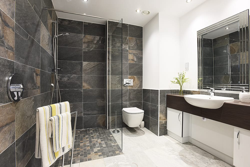 A modern bathroom featuring dark slate-effect wall tiles and a glass walk-in shower with a stone-pebble floor. The room includes a white wall-mounted toilet, a large vanity with a white vessel sink, and a rectangular mirror with a decorative frame.