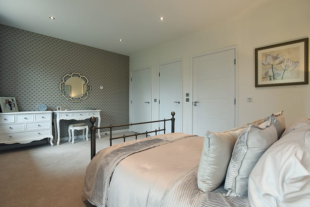 An elegant bedroom featuring a black metal bed frame with neutral-toned bedding and a grey patterned accent wall. To the left, there is a white vintage-style dressing table and matching chest of drawers, while the right side shows white doors and a framed piece of artwork.