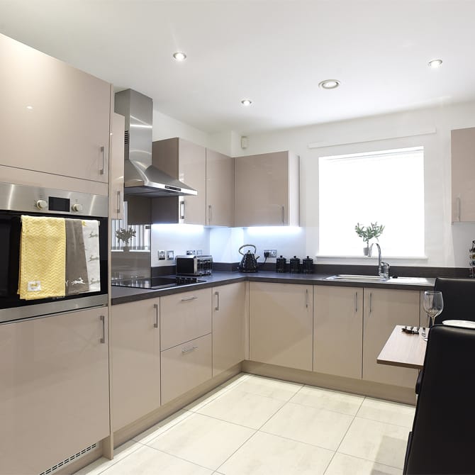 Modern fitted kitchen with beige cabinets, integrated oven and stainless steel extractor hood, dark worktops, under-cabinet lighting, and a bright window above the sink.