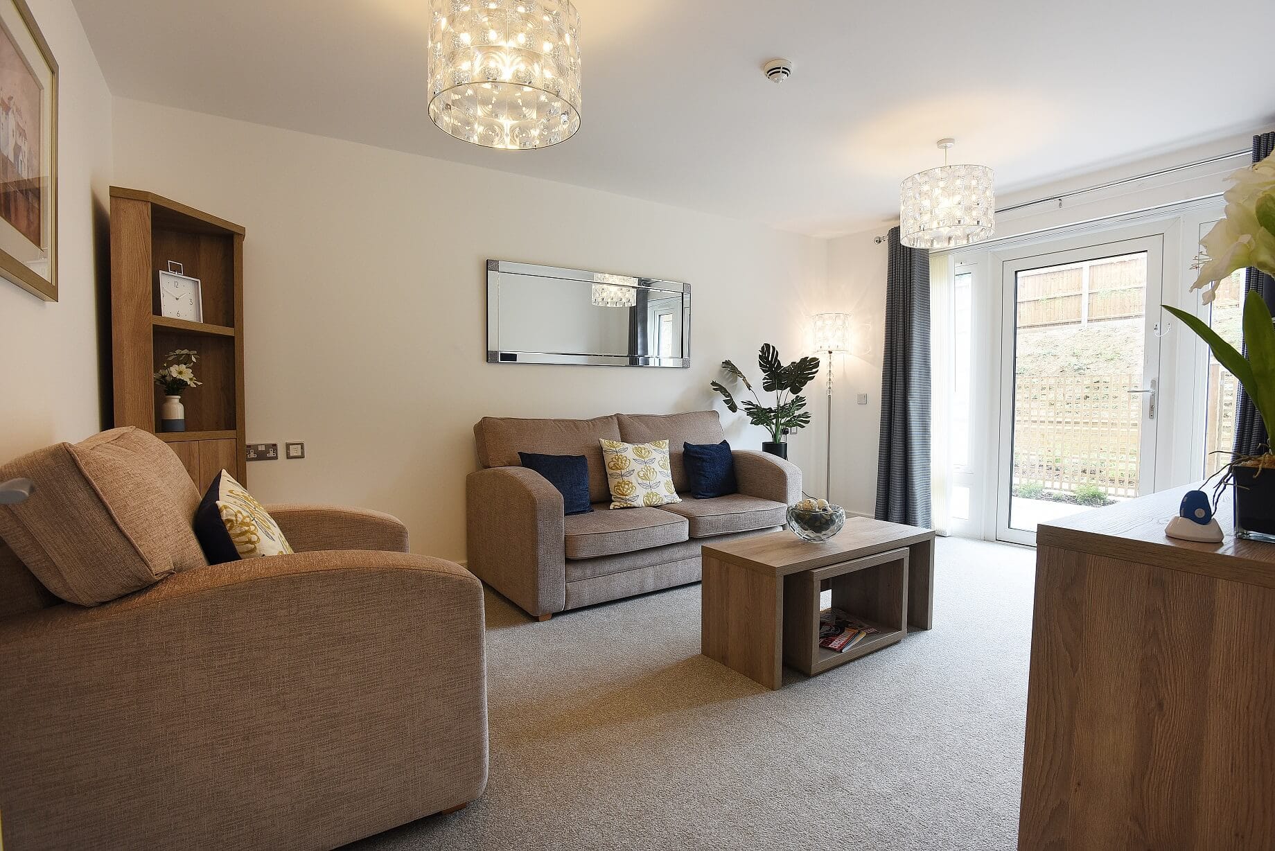 A modern living room with neutral-toned furniture, featuring a beige sofa and matching armchair, a light wood coffee table, and a horizontal mirror on a white wall. Two crystal-style pendant lights hang from the ceiling, and glass patio doors lead to an outdoor area.