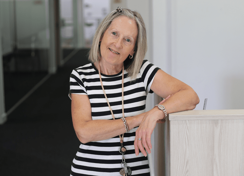 Smiling woman with short light hair wearing a black-and-white striped dress, leaning on a light wooden counter in a modern office setting.