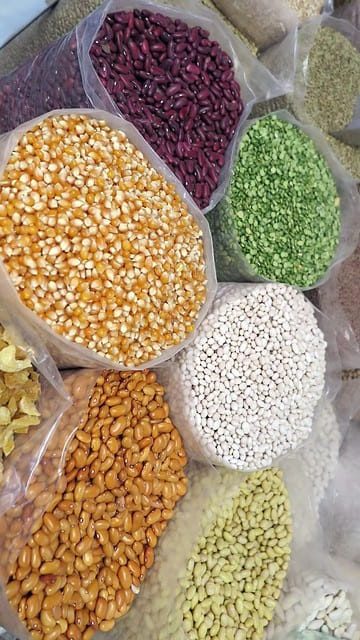 Assorted dried grains and beans displayed in open plastic bags, including corn kernels, red beans, lentils, and split peas.