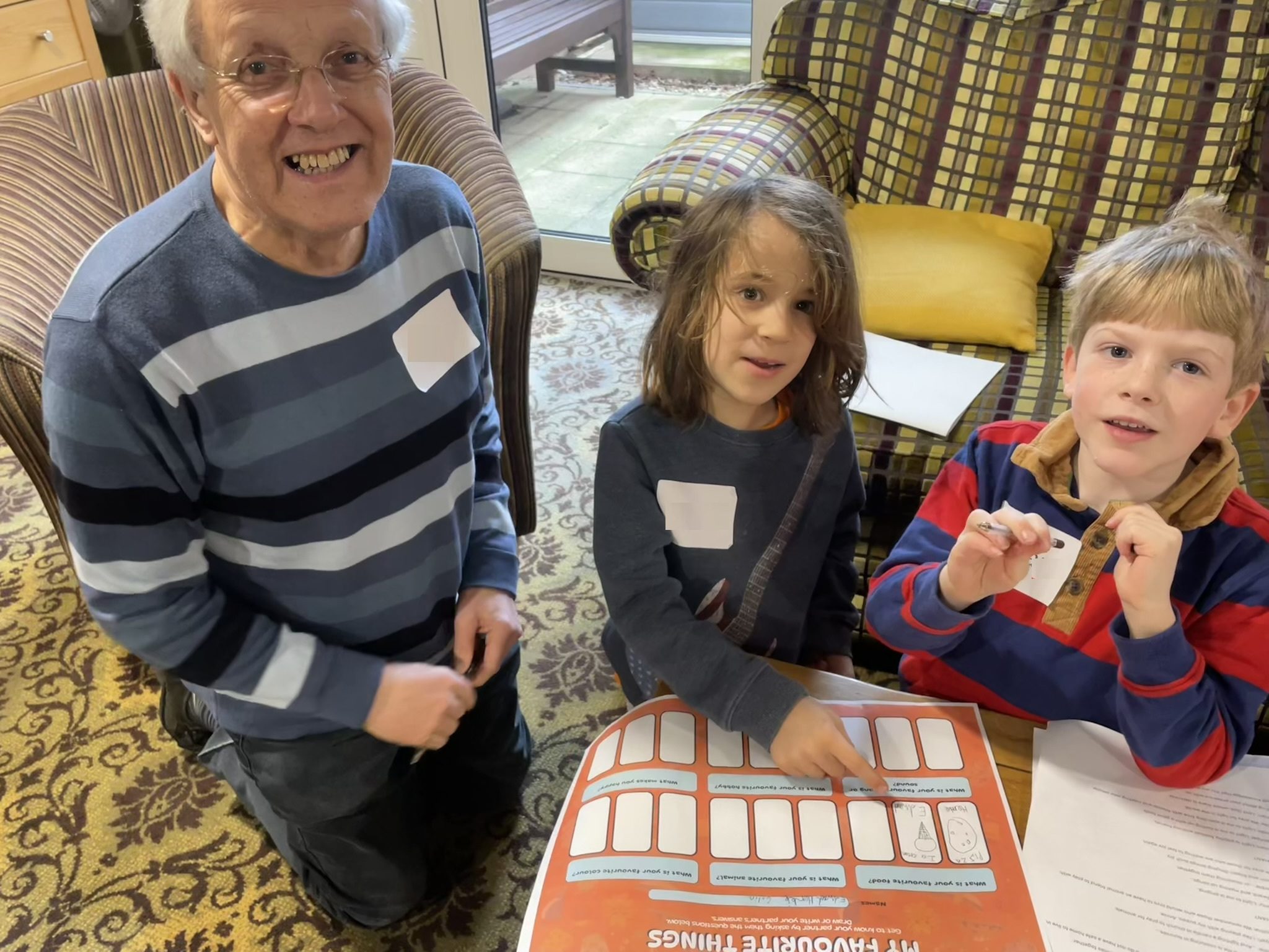 Two children and an older woman sit in the retirement village smiling over a paper exercise.