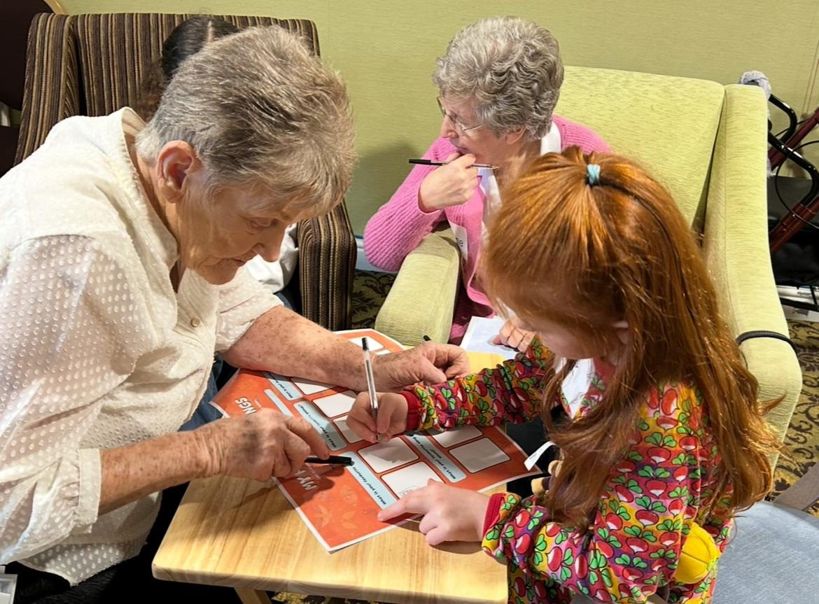 Two older ladies fill in a piece of card with a young redheaded girl.