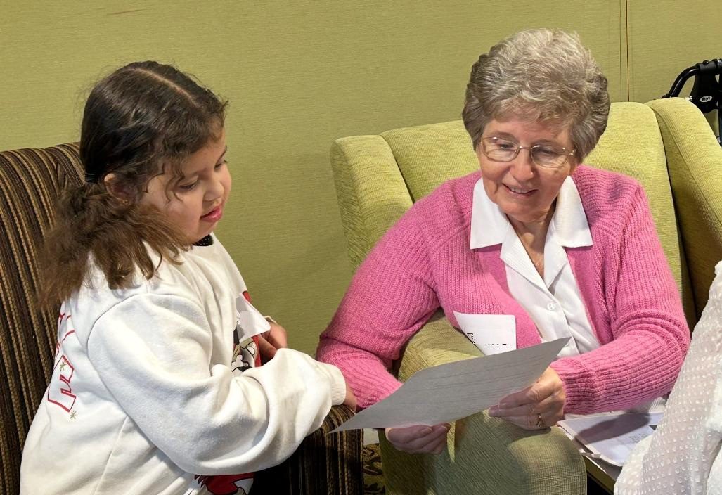 An older woman in a pink sweater reads a piece of card with a young girl in a jumper and pigtails.